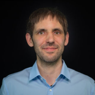Portrait-Bild von David Tanzer, der ein blaues Hemd trägt, auf schwarzem Hintergrund